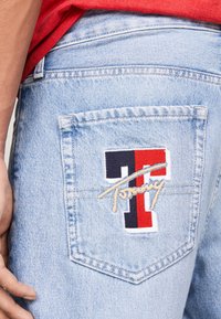 Lyseblå denimjeans med en baklomme som har en flerfarget brodert logo, inkludert røde, blå og hvite detaljer.