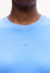 Jasnoniebieska koszulka z krótkim rękawem z bawełny, z prążkowaną okrągłą dekoltą; z białym logo "TOMMY JEANS" oraz małym akcentem w kolorze czerwonym, białym i niebieskim.