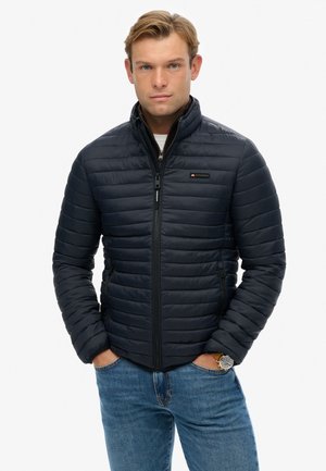 Marineblauwe pufferjack met horizontaal gestikte patronen, hoge kraag en een ritssluiting aan de voorkant. Gepromoot met blauwe jeans en een horloge.