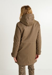 Abrigo largo marrón con capucha, de textura suave y detalles de costura. Llevado sobre pantalones con un estampado de leopardo.