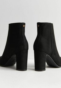 Bottines en daim noir avec un talon épais, présentant une texture lisse et un design minimaliste, rehaussé de petits accents métalliques.
