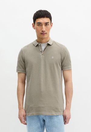 Mężczyzna w beżowej koszulce polo na krótki rękaw, pod którą ma białą podkoszulkę, oraz w jasnoniebieskich dżinsach, stoi na tle jednolitego białego tła.