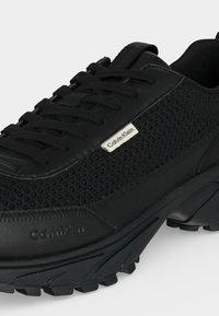 Μαύρο αθλητικό παπούτσι Calvin Klein με υφασμάτινη υφή, δέσιμο με κορδόνια μπροστά και χοντρή λαστιχένια σόλα σχεδιασμένη για κράτημα σε εξωτερικούς χώρους.