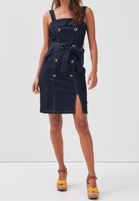 Džínové šaty s přiléhavým střihem, tmavým praním, předním rozparkem, zlatými knoflíky a páskem v pase. Mají široké ramenní popruhy a kontrastní švy.