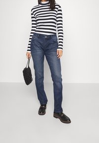 Langarmoberteil mit schwarz-weißen Streifen, kombiniert mit dunkelblauen Jeans. Das Modell hält eine schwarze geflochtene Handtasche und trägt schwarze Schuhe mit einer Schnallendetail.