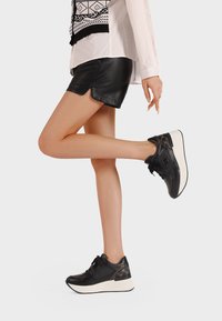 Zapatillas negras con parte superior de cuero brillante, acentos de tela estampada y suela de cuña blanca. Combinadas con pantalones cortos de cuero negros.