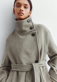 Kvinna i en högkragad grå ullrock med knappar och midjebälte, stående mot en enkel vit bakgrund.