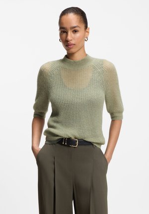 Femme portant un pull en maille vert clair à manches courtes et un pantalon olive foncé avec une ceinture noire, debout les mains dans les poches sur fond blanc.