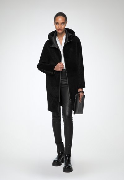 Cappotto in pelliccia sintetica nera con cappuccio, dotato di zip frontale, indossato sopra pantaloni in pelle neri e stivali neri a suola spessa, tenuto con una borsa a mano nera.