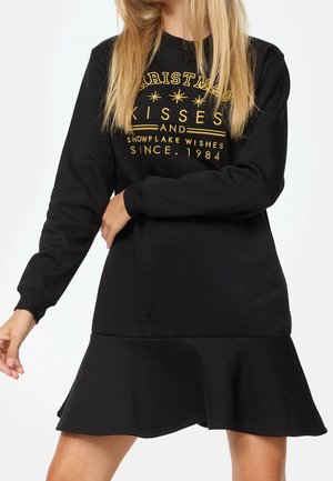 Femme portant une robe noire à manches longues avec un texte doré sur la poitrine indiquant « Christmas kisses and snowflake wishes since 1984 ».