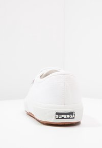 Zapatilla de lona blanca con puntera redonda, suela de goma texturizada y logo negro "SUPERGA" en el talón. Diseño simple y minimalista.