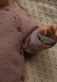 Pleteni sivkini kardigan z lesenimi gumbi, z rebrastim ovratnikom in teksturiranim vzorcem, nošen čez belo dolgoročno majico.