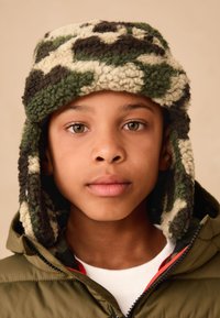 Green and brown camouflage fur hat with ear flaps, soft texture, worn with an olive-green zip-up jacket and a white collar.