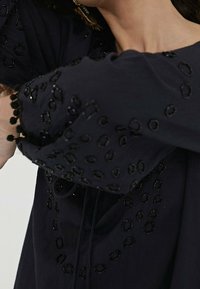 Marinblus med intrikata svarta cirkulära broderier och texturerade ärmar, med pom-pom detaljer vid ärmsluten och en avslappnad passform.