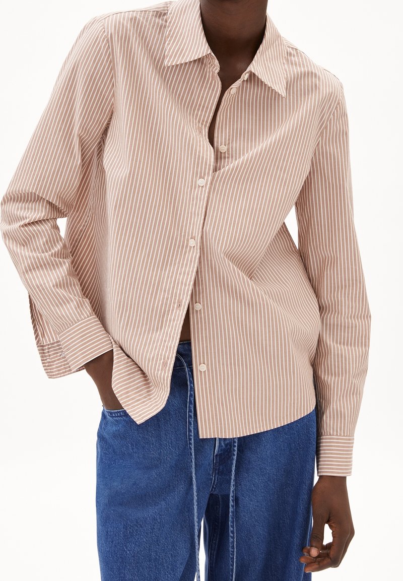 Personne portant une chemise beige et blanche à rayures verticales, boutonnée, à manches longues, et un jean en denim bleu, main dans la poche, sur fond blanc.