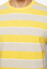 Camiseta a rayas en colores amarillo, gris claro y blanco. Tejido ligero con cuello redondo y mangas cortas.