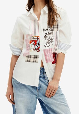 Kobieta w białej koszuli na guziki z podwiniętymi rękawami i naszywkami z postacią Myszki Miki, w połączeniu z jasnoniebieskimi dżinsami.