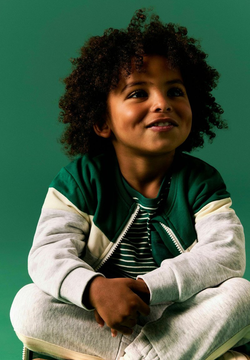 Jeune enfant aux cheveux bouclés, portant une veste verte et blanche et une chemise rayée, assis en tailleur sur un fond vert.
