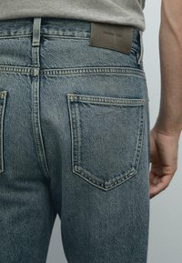 Blue denim jeans with a faded finish, featuring two back pockets, visible stitching, and a brown leather label at the waistband.