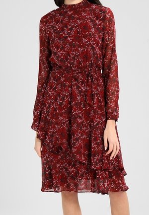Femme portant une robe rouge à fleurs, manches longues, longueur genoux, avec un col montant et une ceinture nouée à la taille.