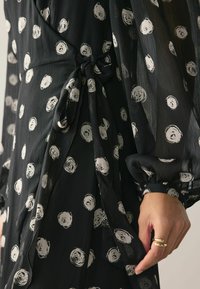 Rochie neagră cu talie legată, fabricată dintr-un material transparent cu modele circulare albe, și cu mâneci lungi voluminoase, cu manșete elastice.