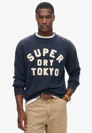 Granatowy sweter z bawełny, z dużym beżowym napisem: "SUPER DRY TOKYO". Żebrowane mankiety i kołnierz, nałożony na biały t-shirt.