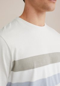 Šviesiai pilkas crewneck trumpomis rankovėmis marškinėliai, papuošti kreminių, smėlio ir švelniai mėlynų horizontalių juostelių. Minkšta, tekstūruota medvilnės medžiaga.