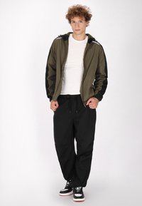 Jachetă verde măsliniu cu mâneci negre, închidere cu fermoar și glugă, asociată cu pantaloni negri, largi, și adidași negri cu accente albe.