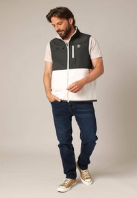 Gilet sans manches pour hommes en vert foncé et blanc, avec fermeture éclair à l'avant, poches et tissu doux. Associé à un jean en denim foncé et des baskets beiges.