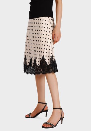 Donna che indossa una gonna beige a pois con bordatura in pizzo nero e sandali con tacco nero a cinturini, in piedi davanti a uno sfondo semplice.