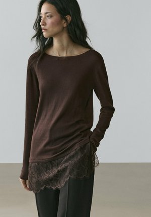 Donna con capelli lunghi e scuri che indossa un top marrone a maniche lunghe con bordo in pizzo, pantaloni neri, guarda verso sinistra su uno sfondo grigio uniforme.