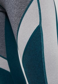 Legíny s vysokým pasem v kombinaci tmavě modrozelené a šedé barvy. Materiál má hladkou texturu s kontrastními švy a geometrickým panelem.