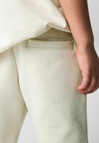 Lys beige sweatpants med struktureret stof, der har en baglomme prægede med "ALLSAINTS." Hånd hviler på lårområdet.