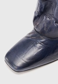 Mornaroblu kožni čizmama sa kvadratnim vrhom i vidljivim nabora, prikazane na bijeloj pozadini.