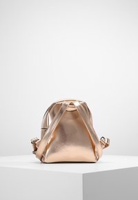 Metallinen ruusukultainen reppu, jossa on ristikkäinen hihnasuunnittelu, sileä pinta, pyöreä muoto ja säädettävät olkahihnat. Kompaktin ja modernin näköinen.