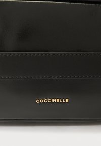 Bolso de mano de cuero negro con textura suave, detalles de costura sutiles y un logo dorado de "Coccinelle". Diseño simple y rectangular.
