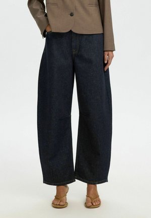 Alakeha inimesest, kes kannab tumedaid laiade säärega teksapükse, beežikat bleiserit ja beežikaid avatud varbaga sandaale valgel taustal.