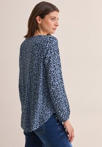 Blaue gemusterte Bluse aus leichtem Stoff, mit langen Ärmeln und gespaltenem Rückensaum. Das Design umfasst abstrakte schwarze Formen auf einem blauen Hintergrund.