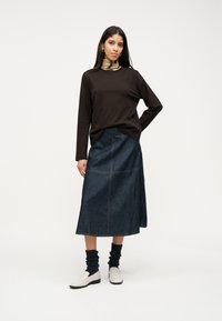 Γυναίκα με μακριά σκούρα μαλλιά, φορώντας μαύρο μακρυμάνικο τοπ, ζιβάγκο με σχέδια, μεσομήκη τζιν φούστα, μαύρες κάλτσες και λευκά μοκασίνια, μπροστά σε λευκό φόντο.