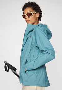 Jachetă de ploaie albastră, ușoară, cu glugă, buzunare cu fermoar și mansete ajustabile. Purtați cu ochelari de soare și având un design elegant.