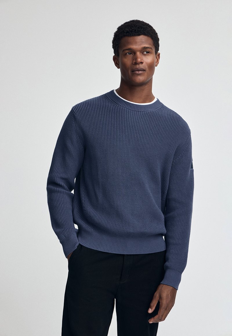 Marineblauwe gebreide trui met een geribde textuur, ronde hals en lange mouwen, gecombineerd met zwarte broeken. Subtiele logo-detail op de mouw.