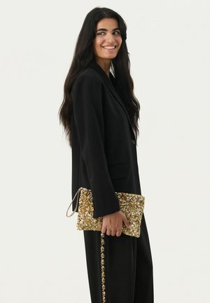 Sacoche recouvert de sequins dorés, de forme rectangulaire, avec une bandoulière marron. Portée avec un blazer noir et un pantalon assorti présentant des accents dorés.