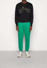 Schwarzer Sweatshirt mit goldener Schrift "grimy", kombiniert mit grünen tapered Sweatpants und hellen Sneakers.