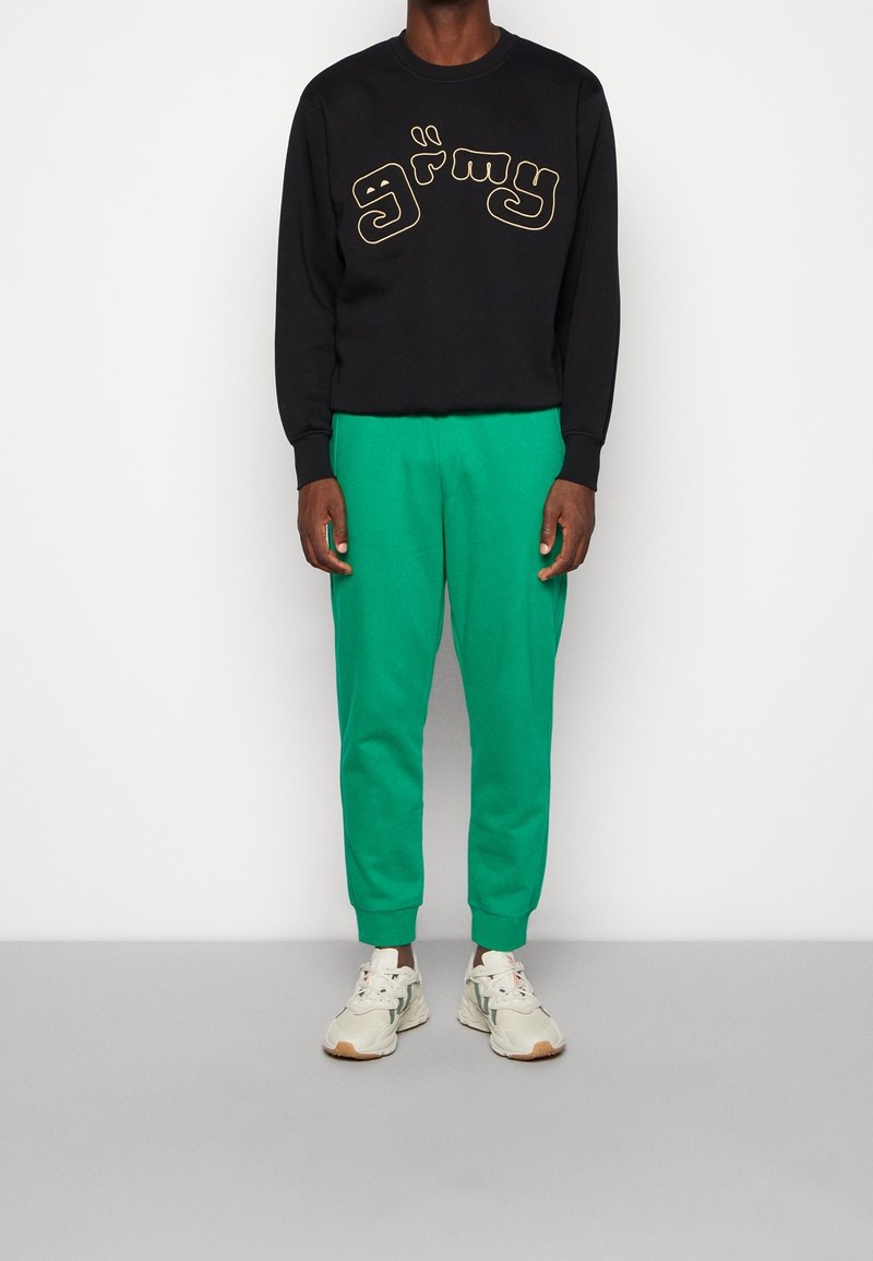 Schwarzer Sweatshirt mit goldener Schrift "grimy", kombiniert mit grünen tapered Sweatpants und hellen Sneakers.