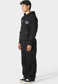 Černá mikina s kapucí a přední kapsou, zdobená vyšitým nášivkem a šňůrkami. V kombinaci s černými tepláky, oba z lehkého materiálu.