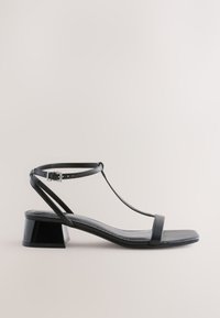 Sandale en cuir noir avec fine bride de cheville, fermeture à boucle, design à bride en T, bout carré et petit talon bloc sur fond neutre.