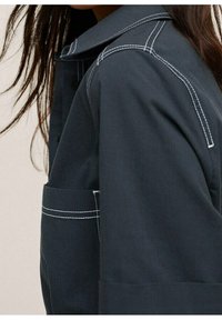 Veste bleu foncé avec surpiqûres blanches, dotée d'un col et d'une poche plaquée sur le devant. Le tissu semble lisse et résistant.