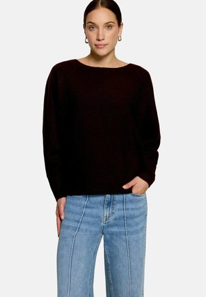 Frau mit dunklen Haaren, die goldene Creolen, einen schwarzen Pullover und hellblaue Jeans mit hoher Taille und sichtbaren Nähten trägt, steht mit der Hand in der Tasche.