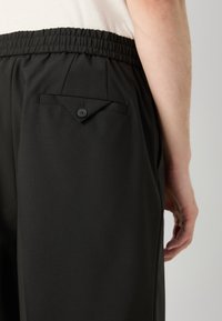 Musta housut, joissa on kerätty joustava vyötärönauha, sileä pinta ja takatasku, jossa on nappikiinnitys ja kolmionmuotoinen laskoskappale.