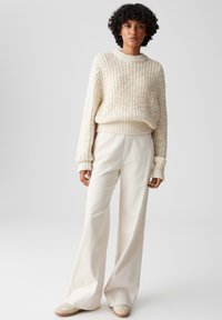 Beiger Strickpullover mit gerippter Textur, kombiniert mit weit geschnittenen beigen Hosen. Das Outfit zeichnet sich durch lockere Passformen und weichen Stoff aus.